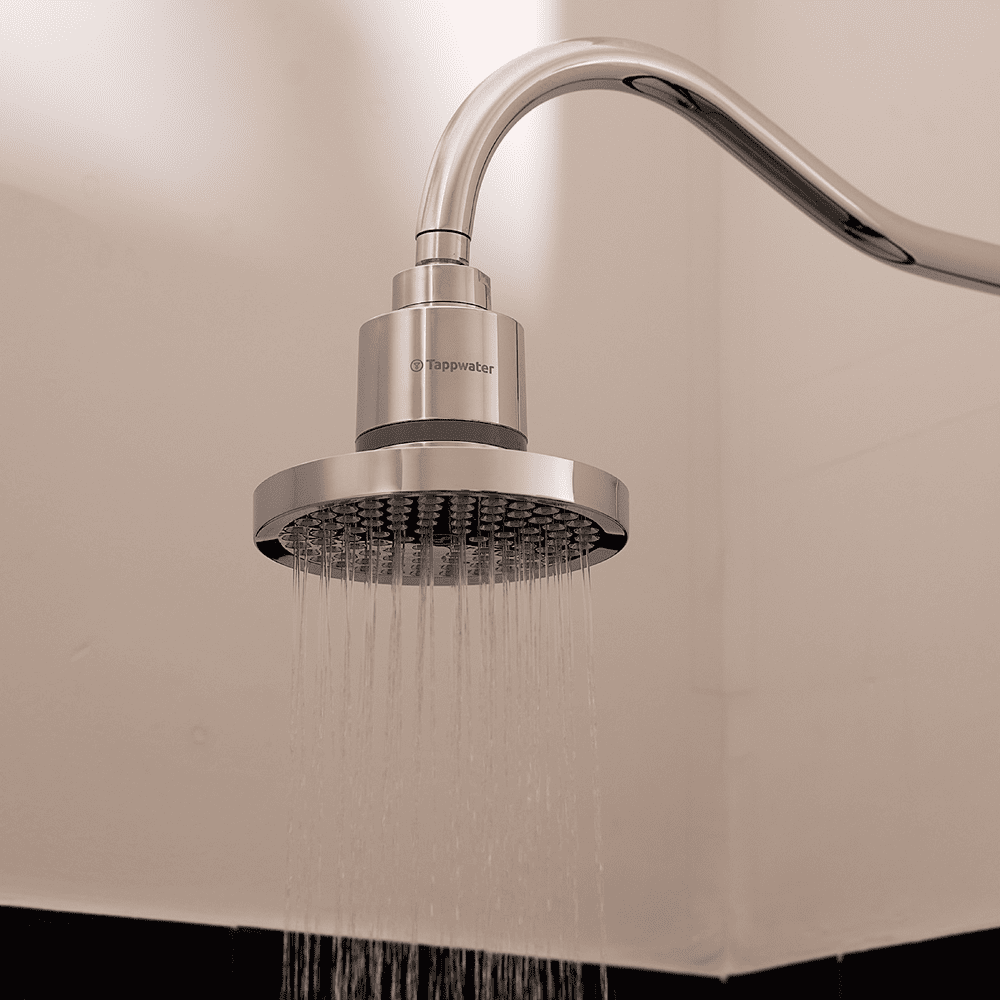 The Tappwater ShowerPro Rain, a modern silver shower head, is mounted on a curved metal pipe and sprays fine streams of water downward against a light-colored bathroom wall.