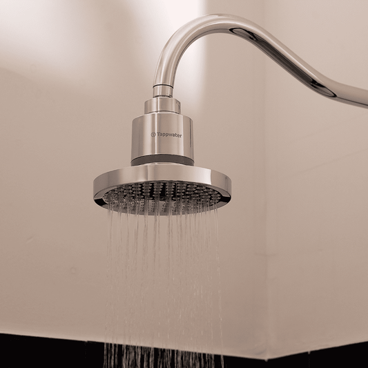 The Tappwater ShowerPro Rain, a modern silver shower head, is mounted on a curved metal pipe and sprays fine streams of water downward against a light-colored bathroom wall.